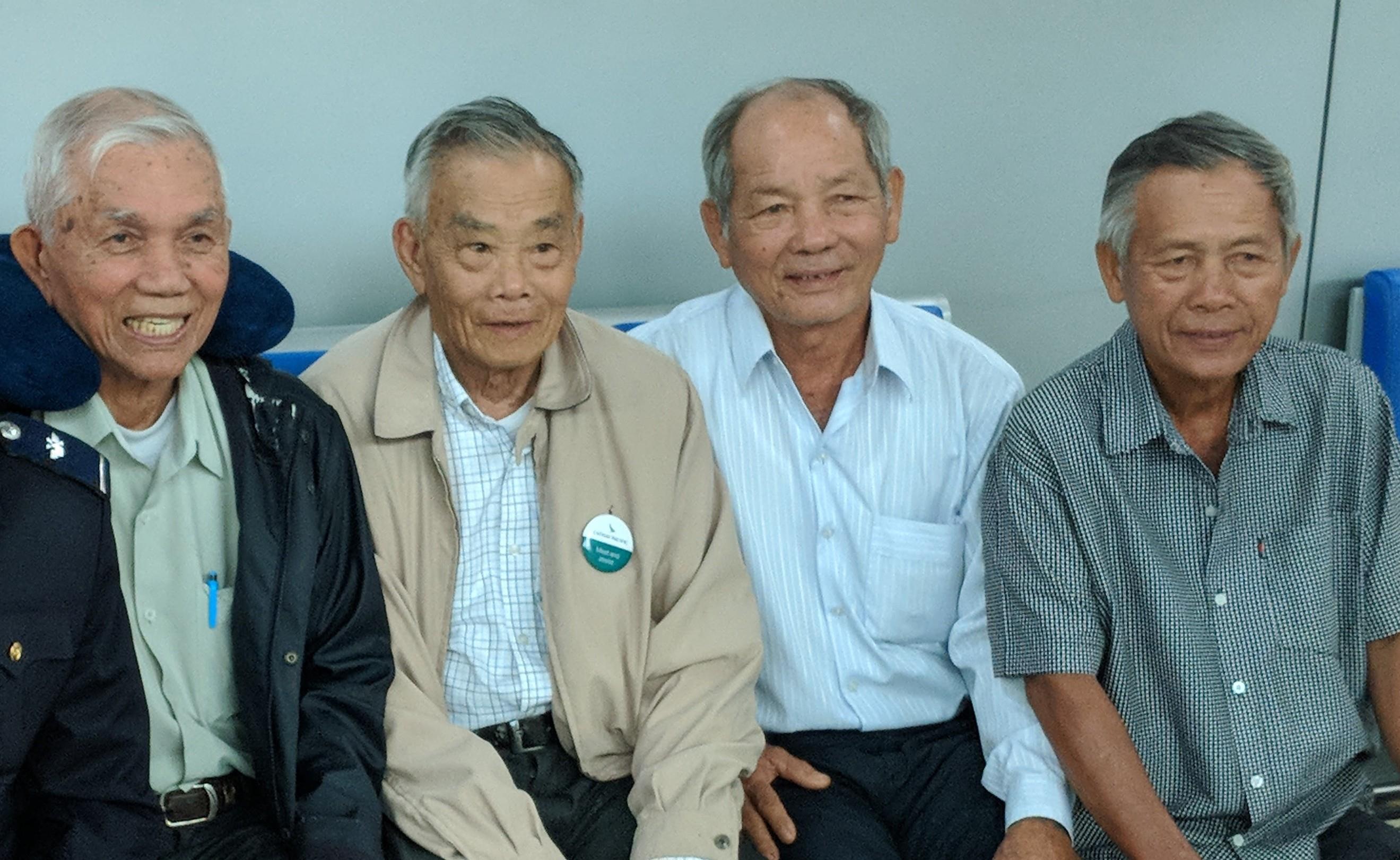 Four elderly men sit close together, smiling and chatting indoors.
