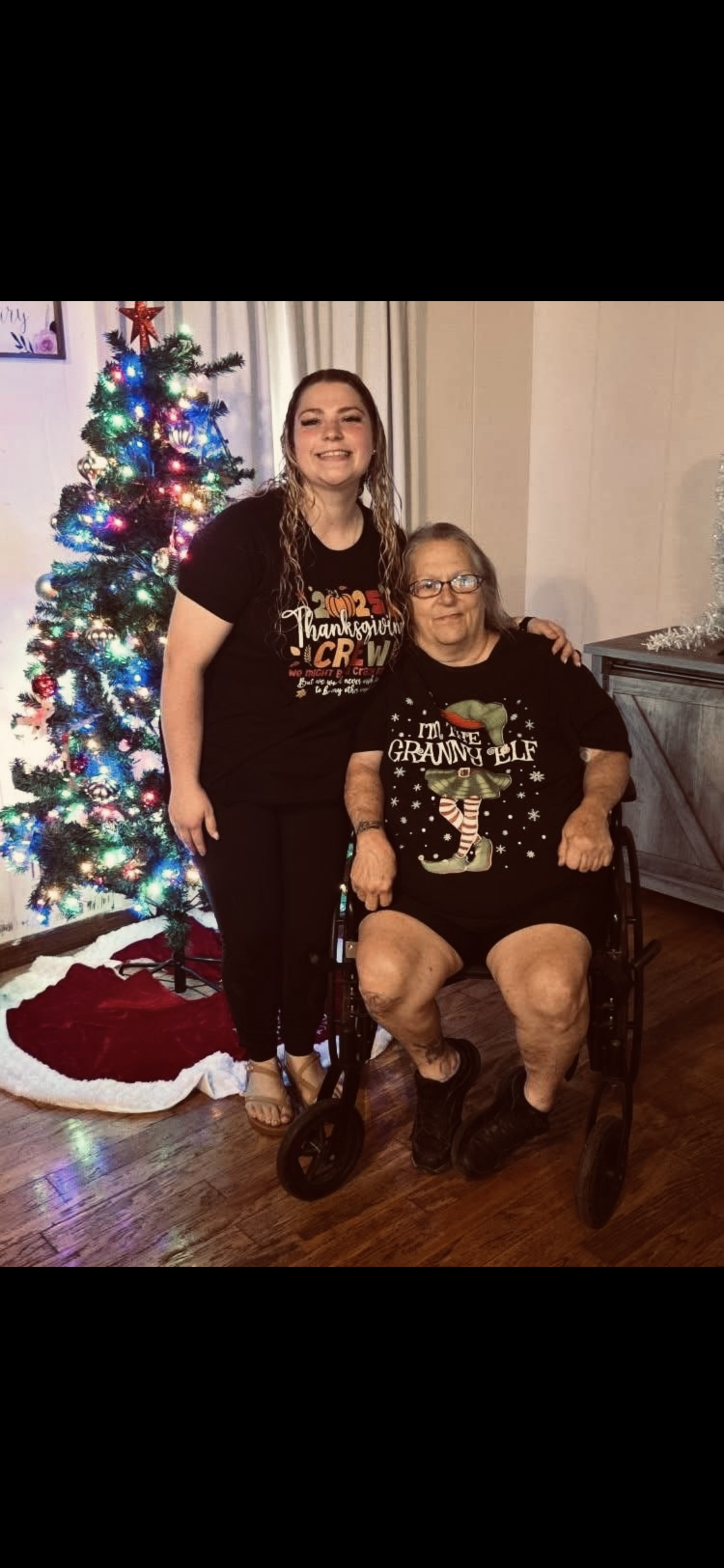 Two family members pose together near a Christmas tree during the holiday season.