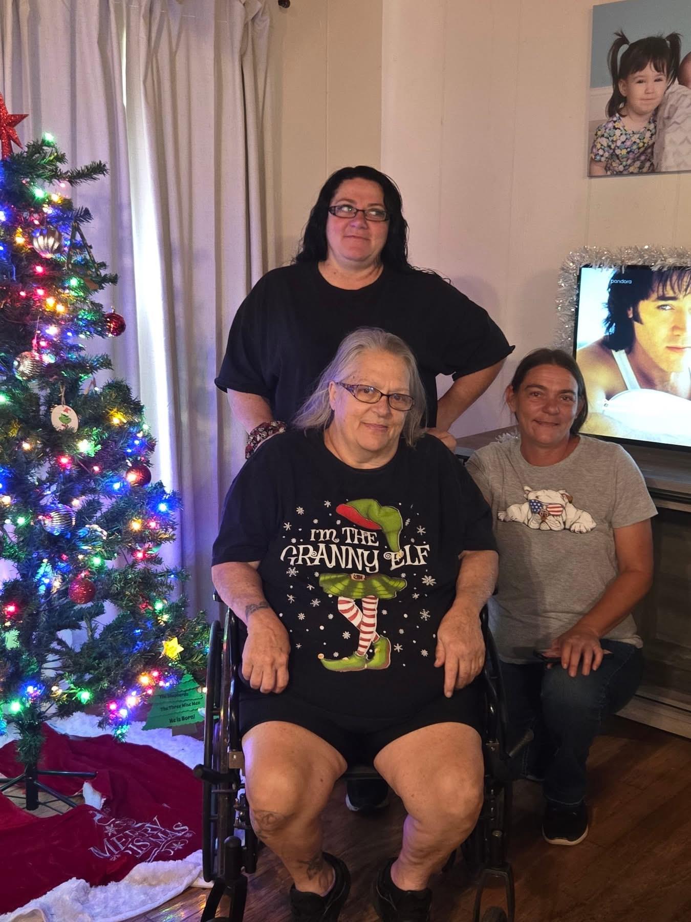 Three family members pose together by a decorated Christmas tree in a cozy living room.