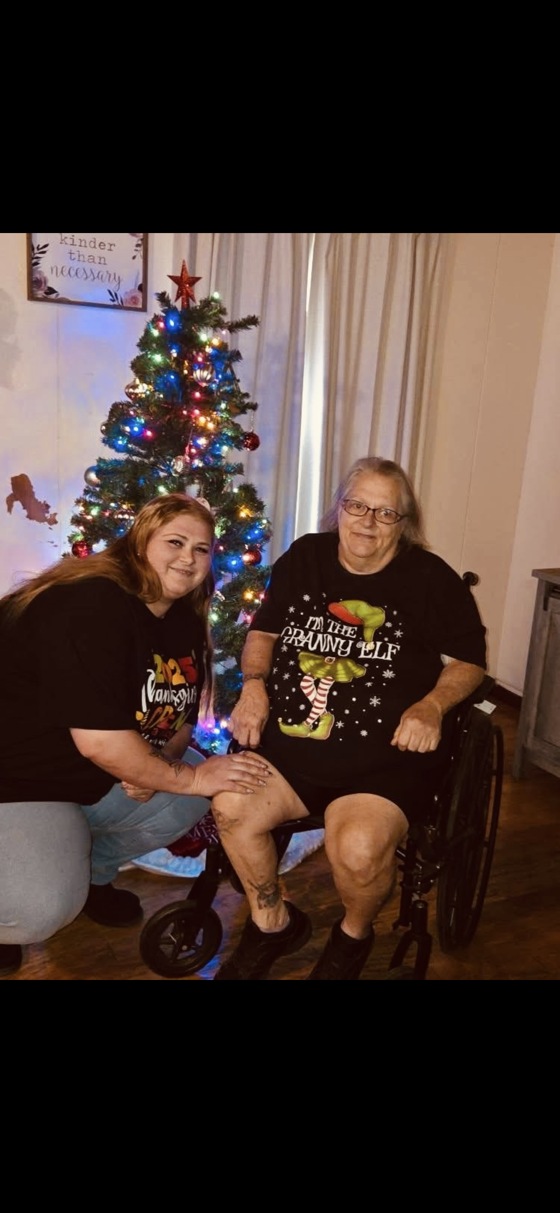 Two women pose by a decorated Christmas tree in a living room. They smile and enjoy the moment.