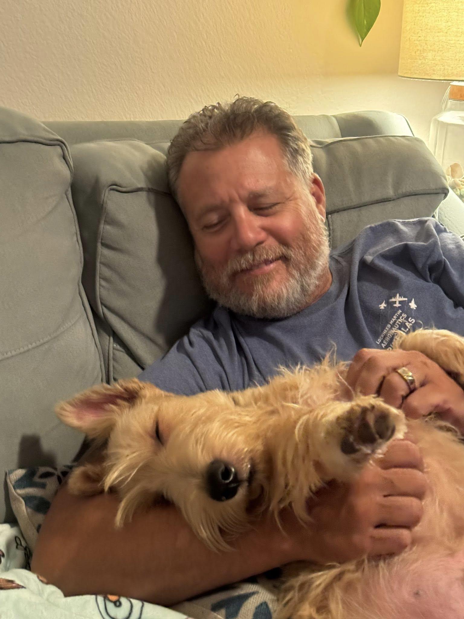 A man relaxes on a couch holding a dog while they share a moment in the living room.
