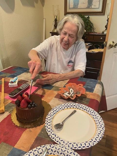 An elderly woman sits at a table and cuts a cake while family members celebrate together.