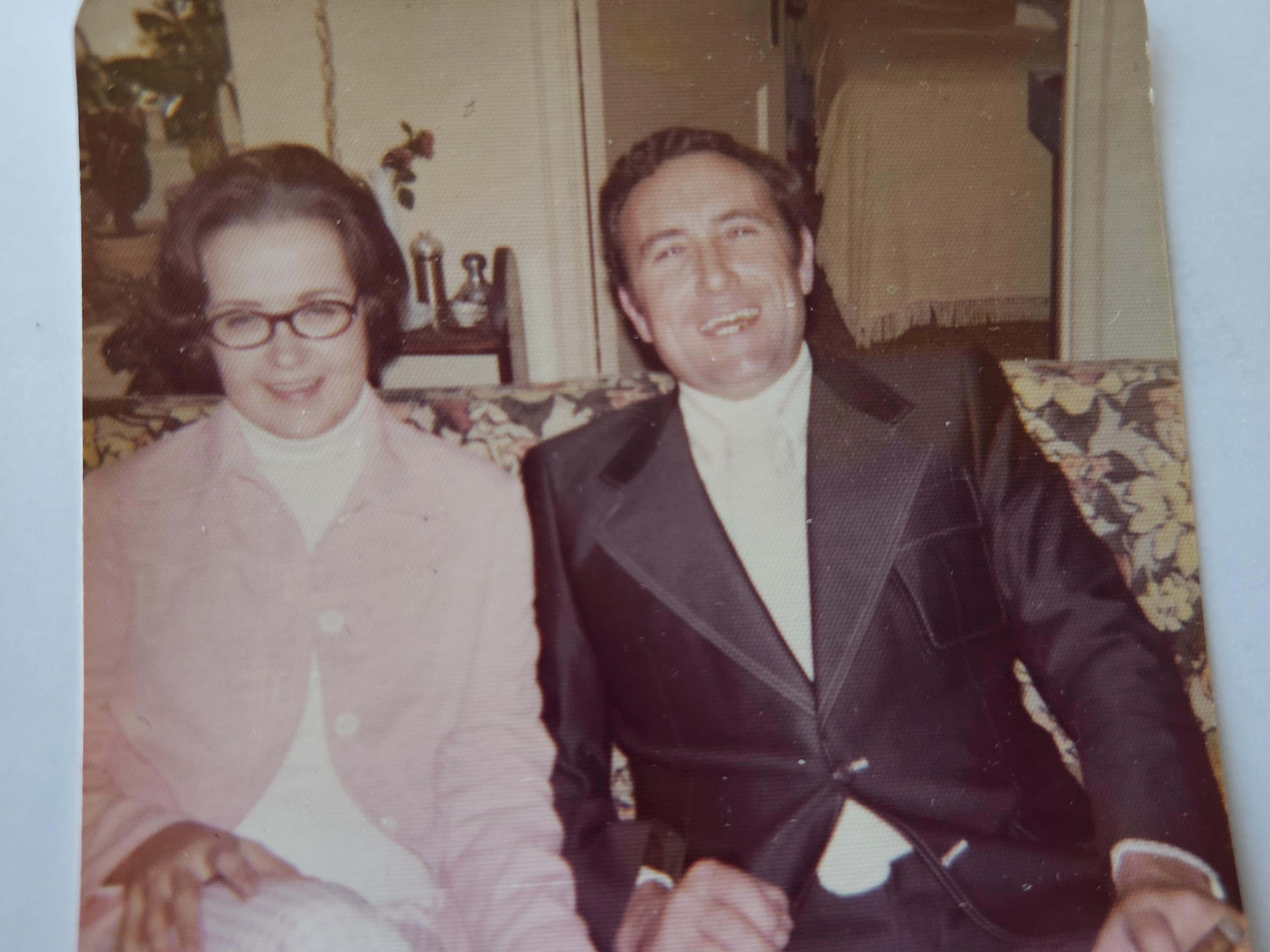 A couple enjoys time together on a couch, smiling at a social event indoors during the evening.