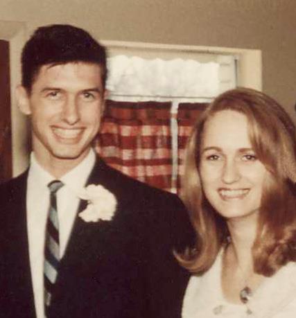 Couple stands close during an event, smiling with formal attire and a floral decoration.