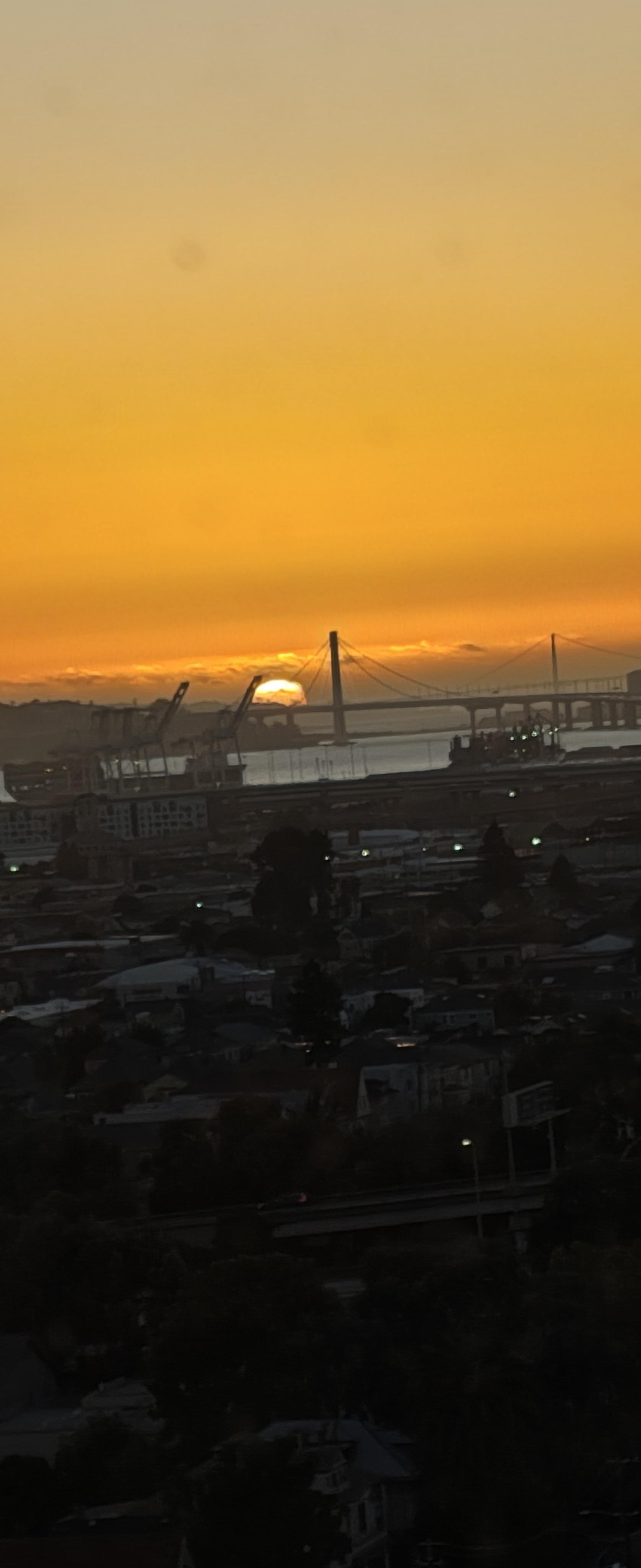 Workers are present at a construction site as the sun sets behind the skyline and bridge.