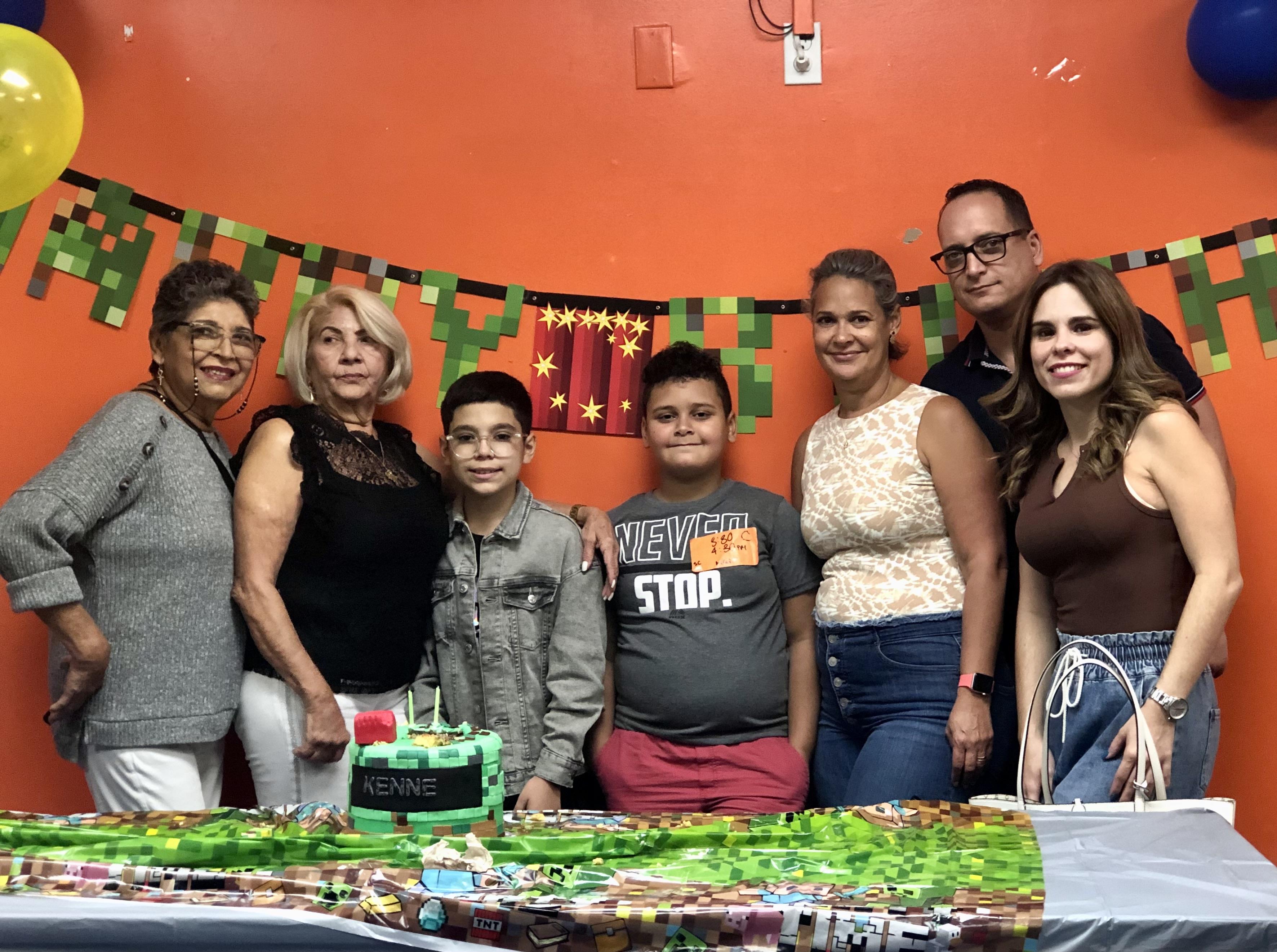 Group of family and friends pose together at a birthday party with cake and colorful decorations.