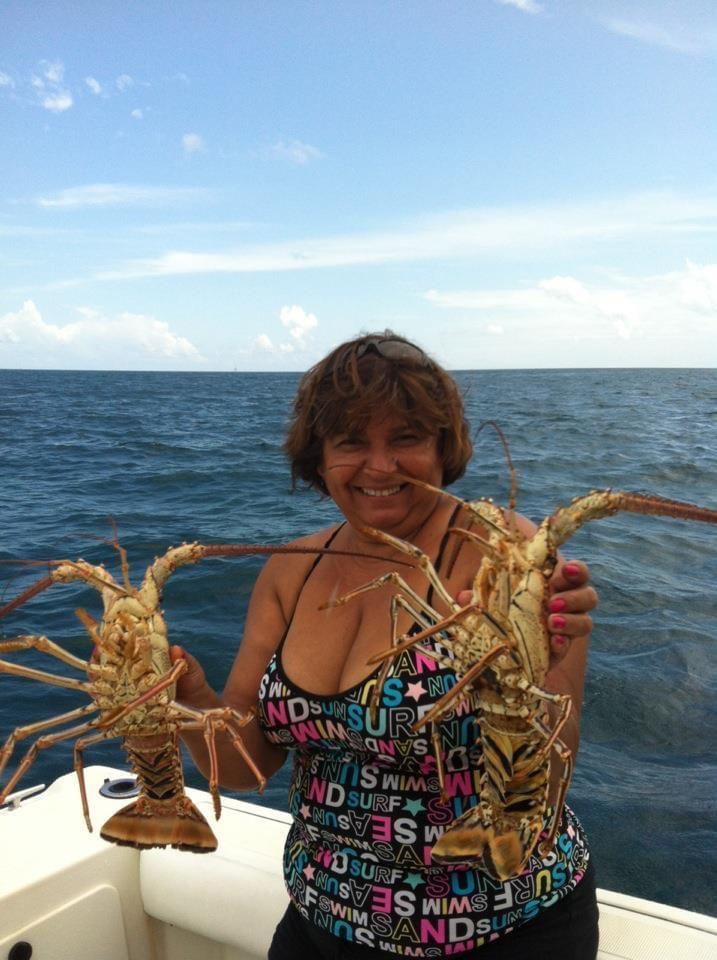A woman smiles while holding two big lobsters on a boat in the ocean under a clear sky.