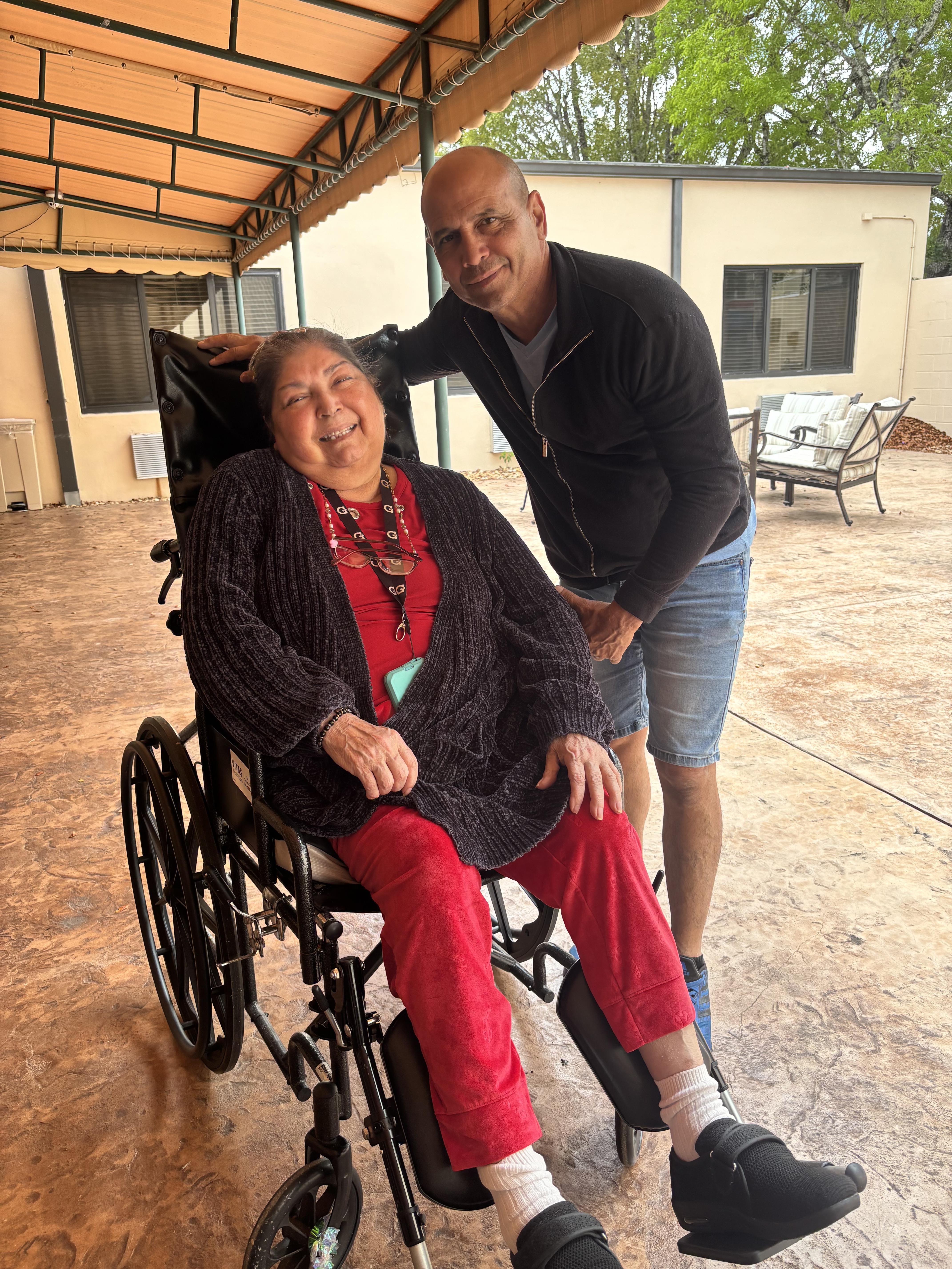 A person is helping a woman in a wheelchair while they smile in front of a building.