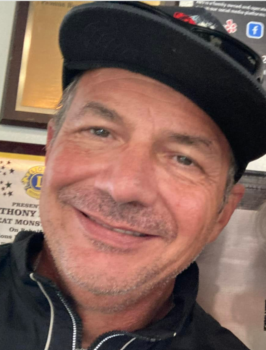 A man stands with a smile in a group setting, enjoying a casual moment with friends.