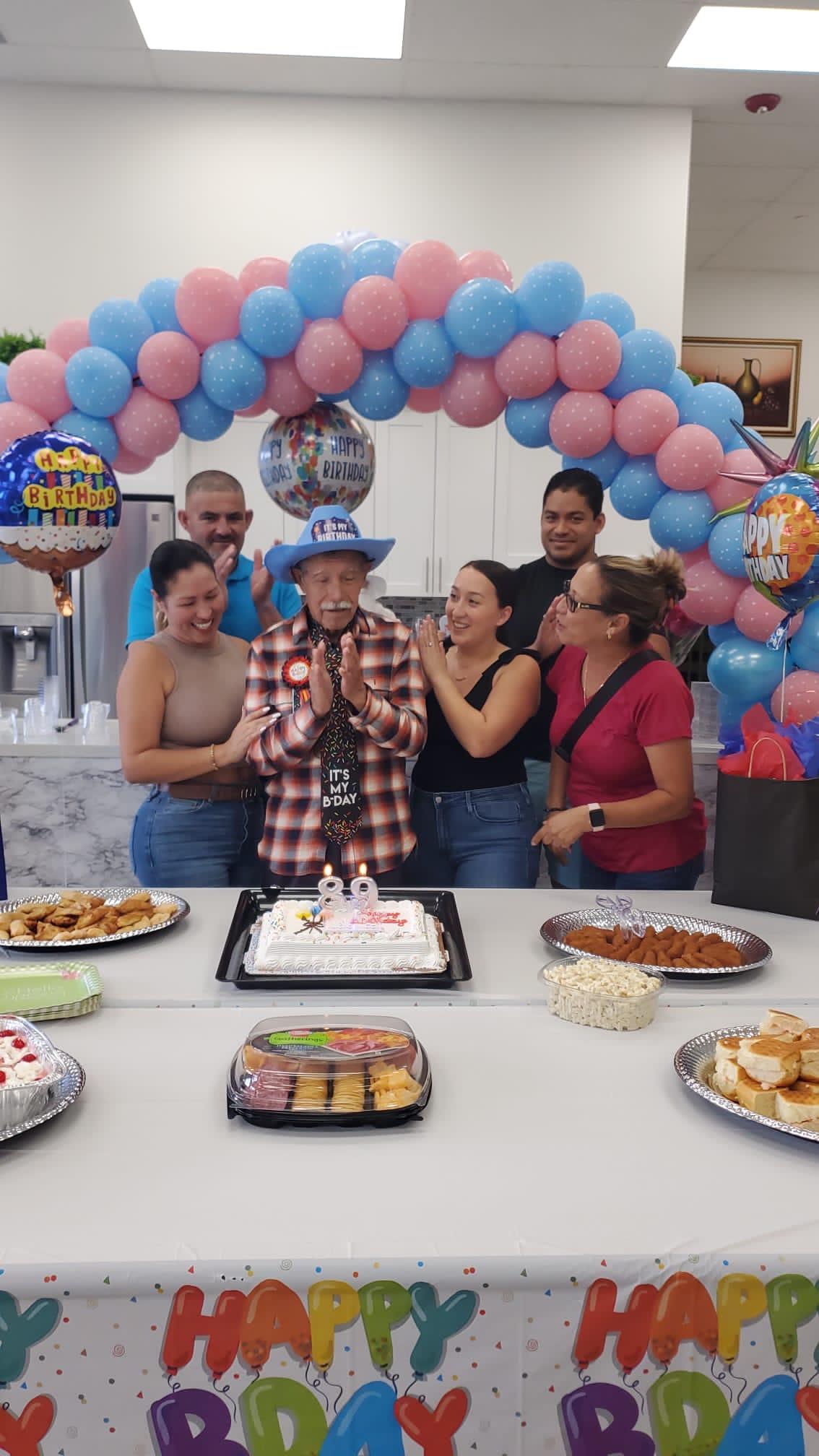 Friends gather to celebrate a birthday with a cake and food while balloons decorate the room.