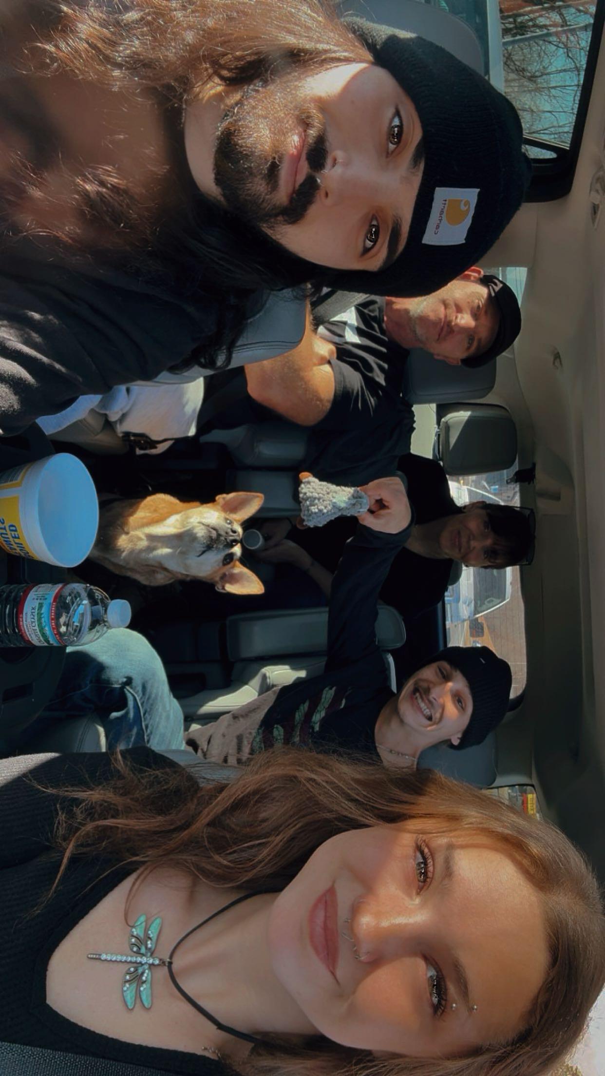 Five friends sit in a car, smiling and having fun while a dog looks out from the back seat.