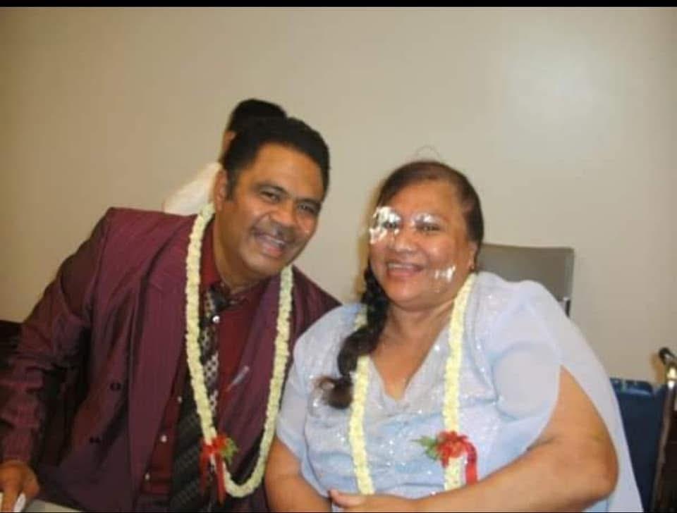 A couple smiles while wearing leis during a family event. They sit close together in a room.