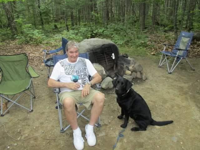 Man enjoys time outdoors with a black dog near a campsite in the forest.
