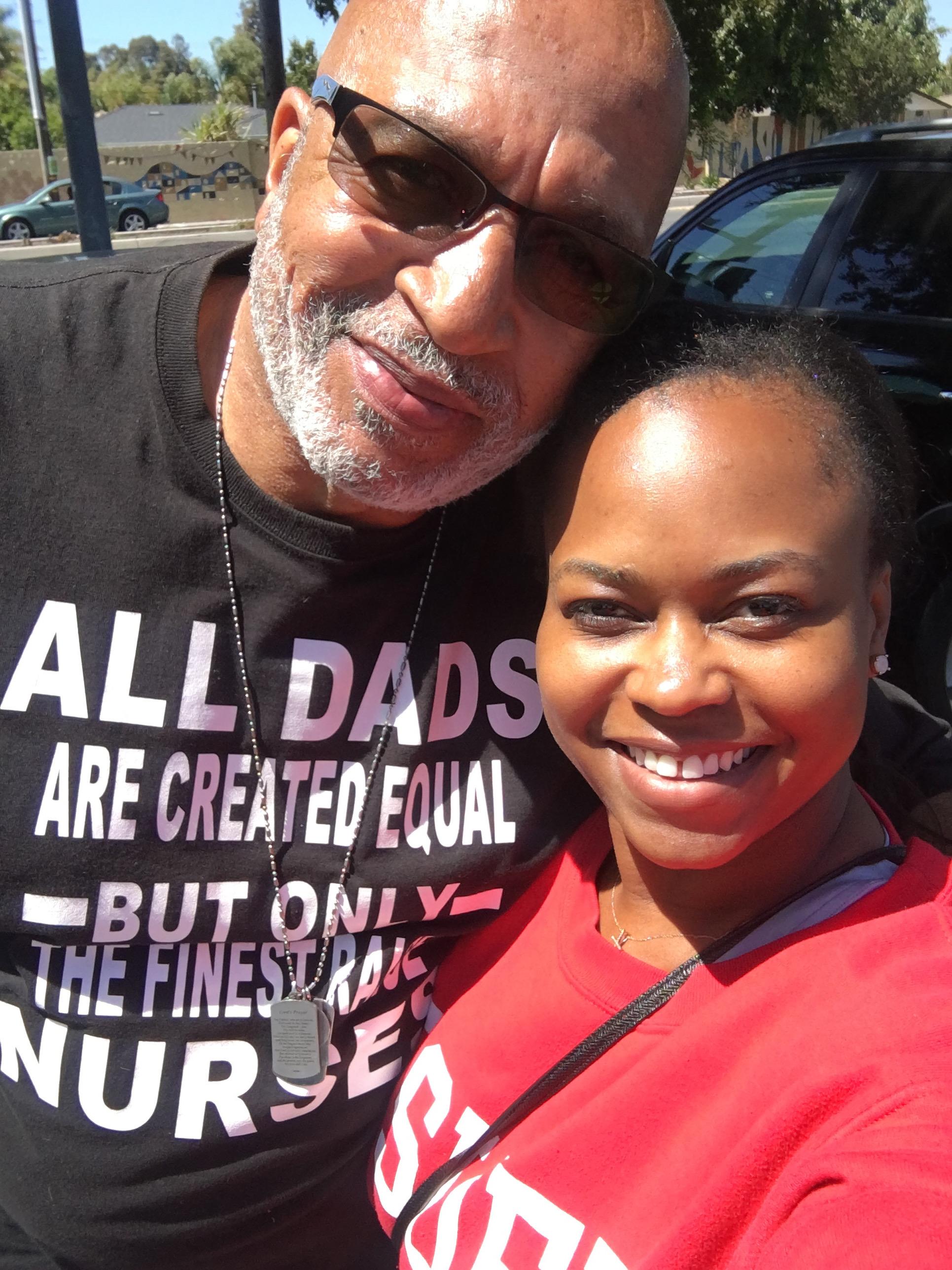 Two individuals take a selfie together with smiles outdoors at a community event under sunlight.