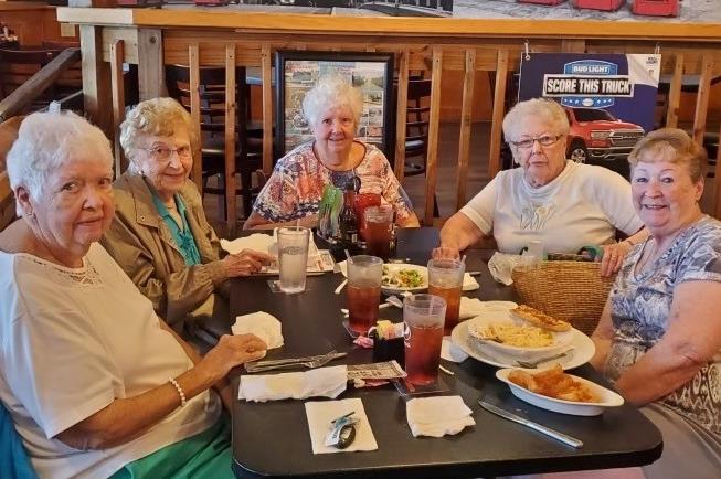 Four women sit at a table, sharing food and drinks while talking and laughing together.