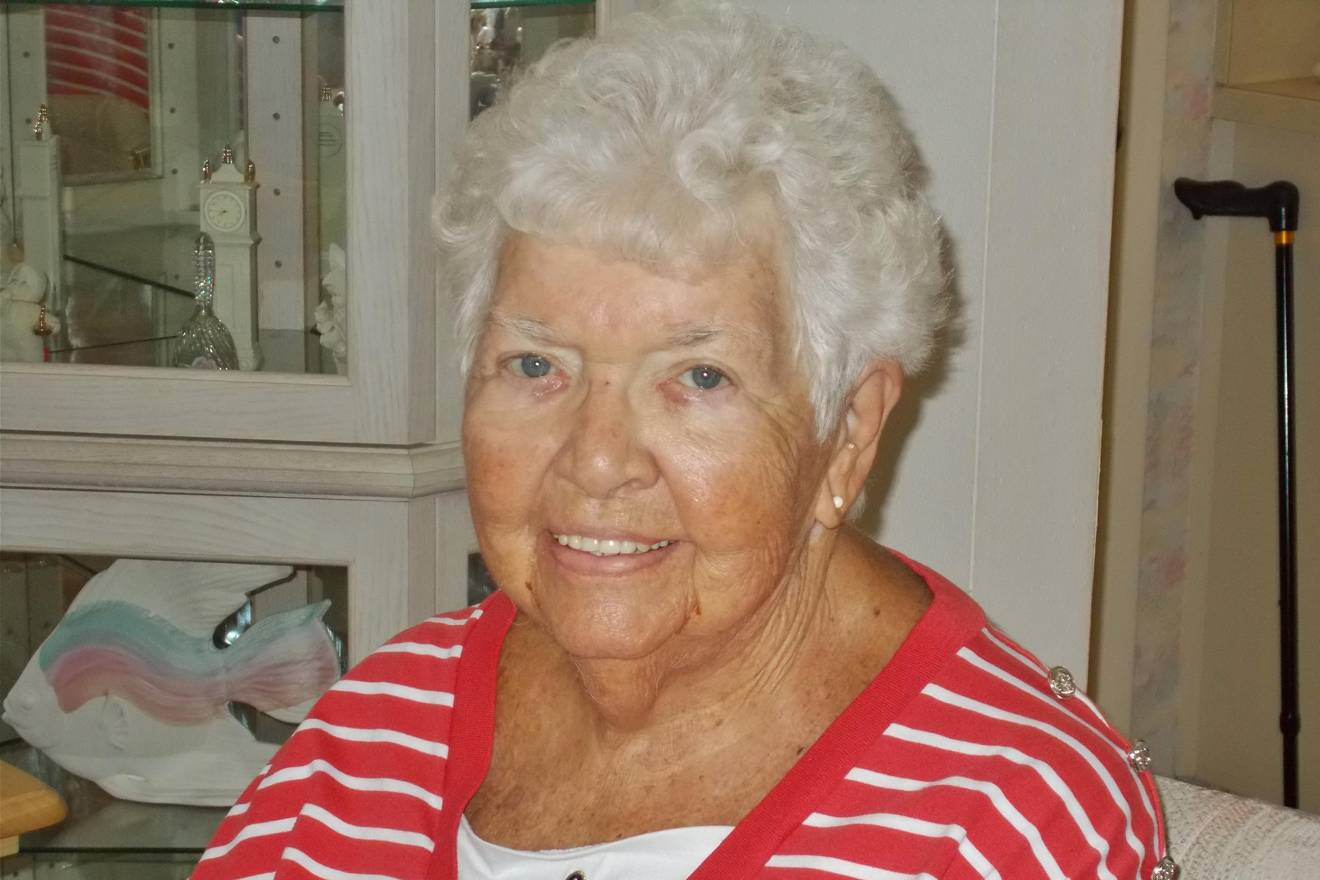 An elderly woman with white hair sits inside a room with a bright smile.