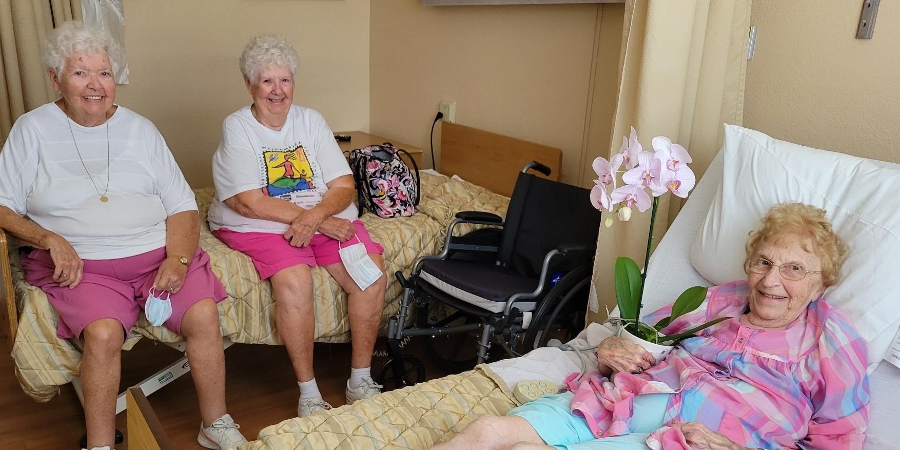 Two women sit on separate beds in a care facility room.