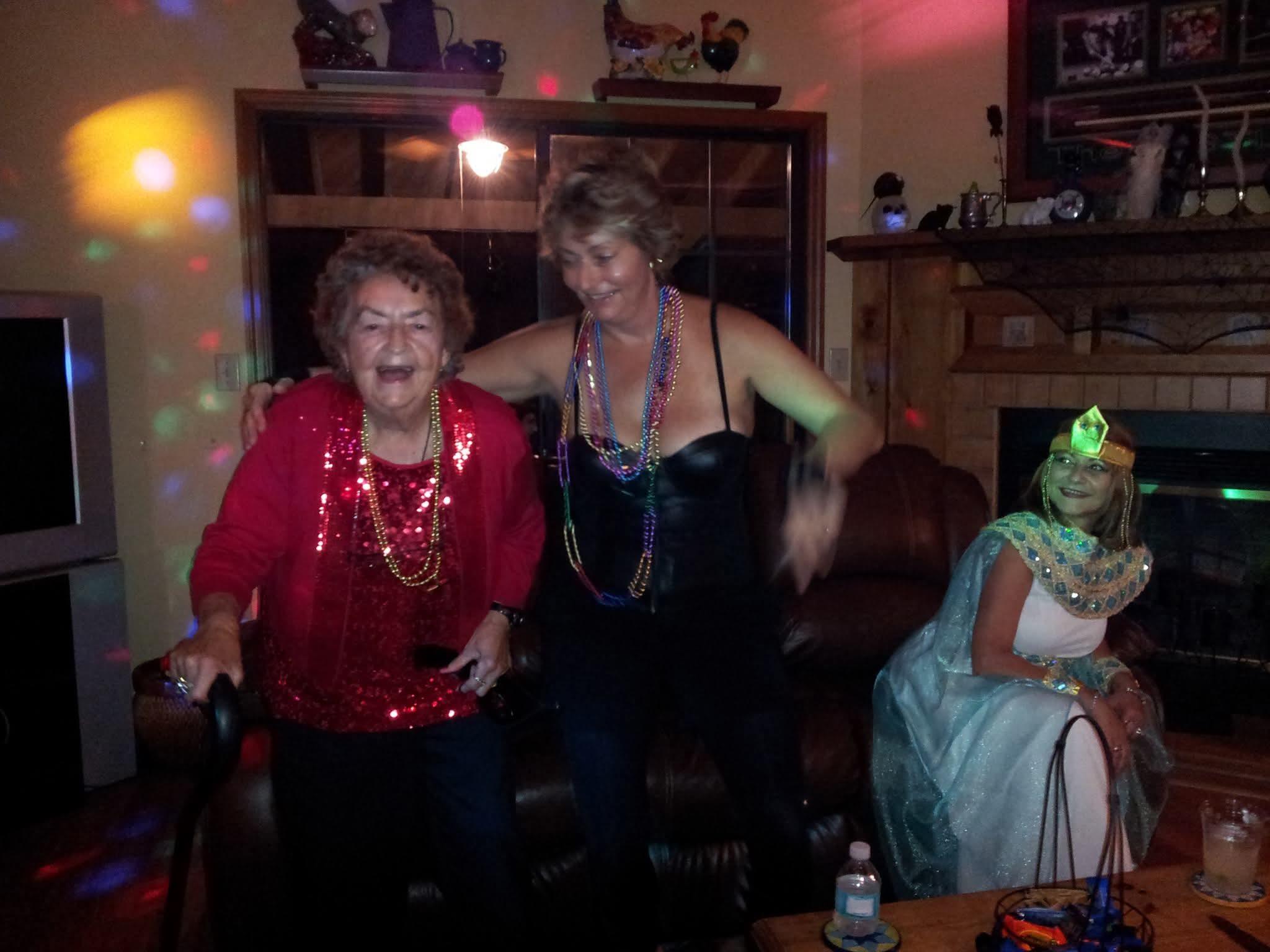 Two women enjoy dancing at a gathering in a lively room while another sits peacefully nearby.