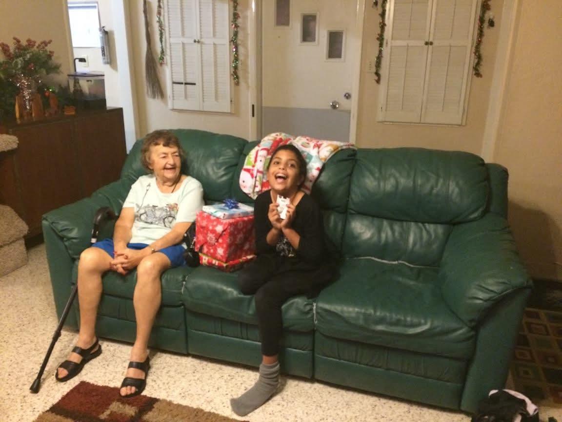 Two children sit on a couch, smiling and excited as they open gifts during a celebration.