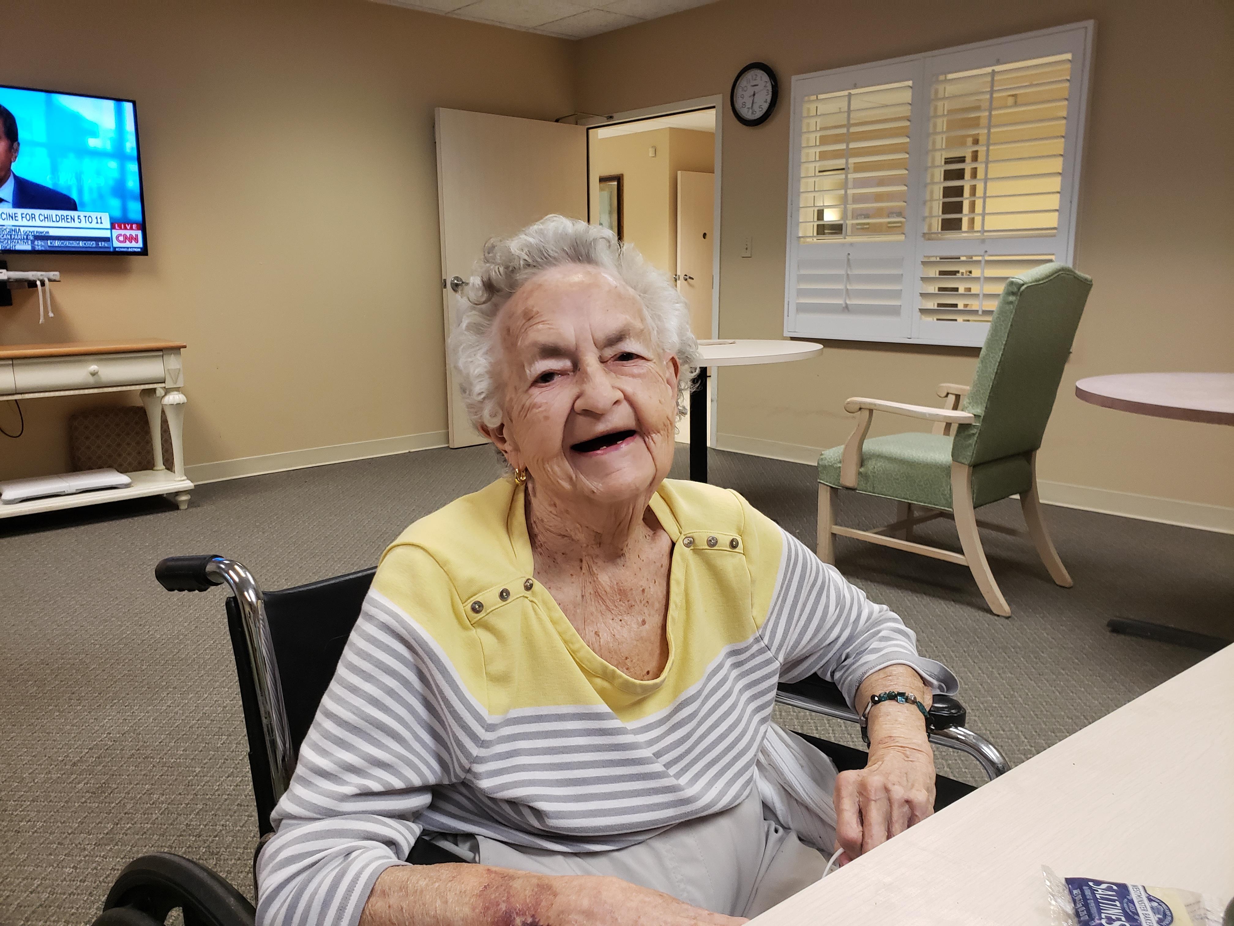 An elderly woman in a wheelchair smiles at the camera in a daytime care facility.