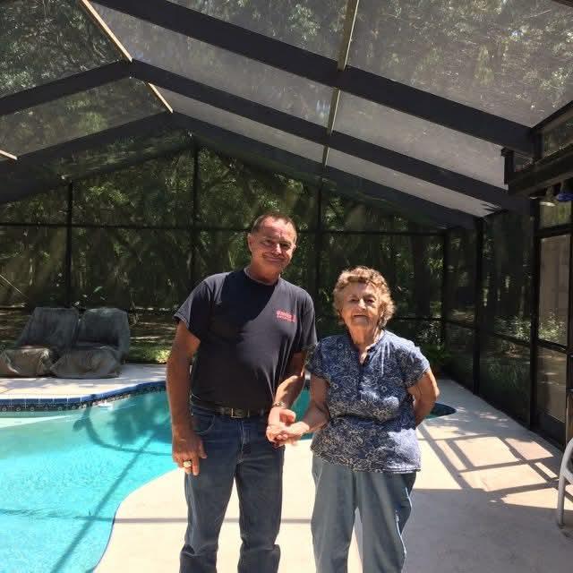 A man and a woman pose for a picture near a swimming pool in a sunny location with a screened patio.
