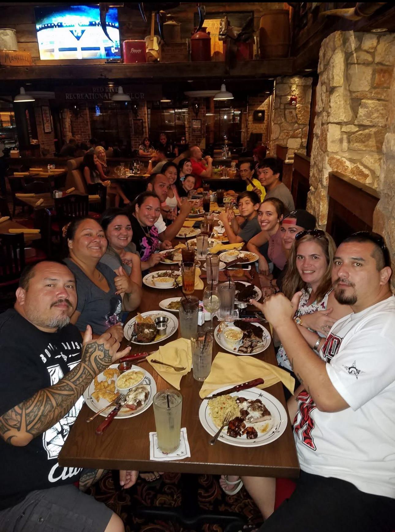 People sit around a long table in a restaurant, sharing food and drinks while smiling.