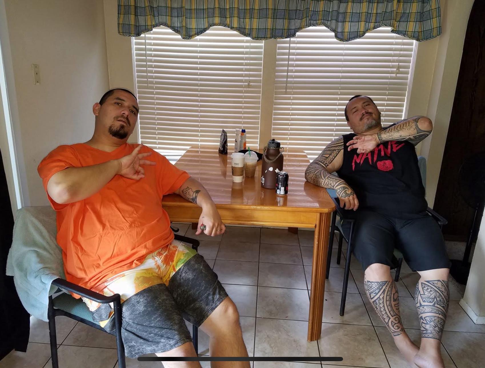 Two men relax in a kitchen, sitting at a table with drinks and food in front of them.