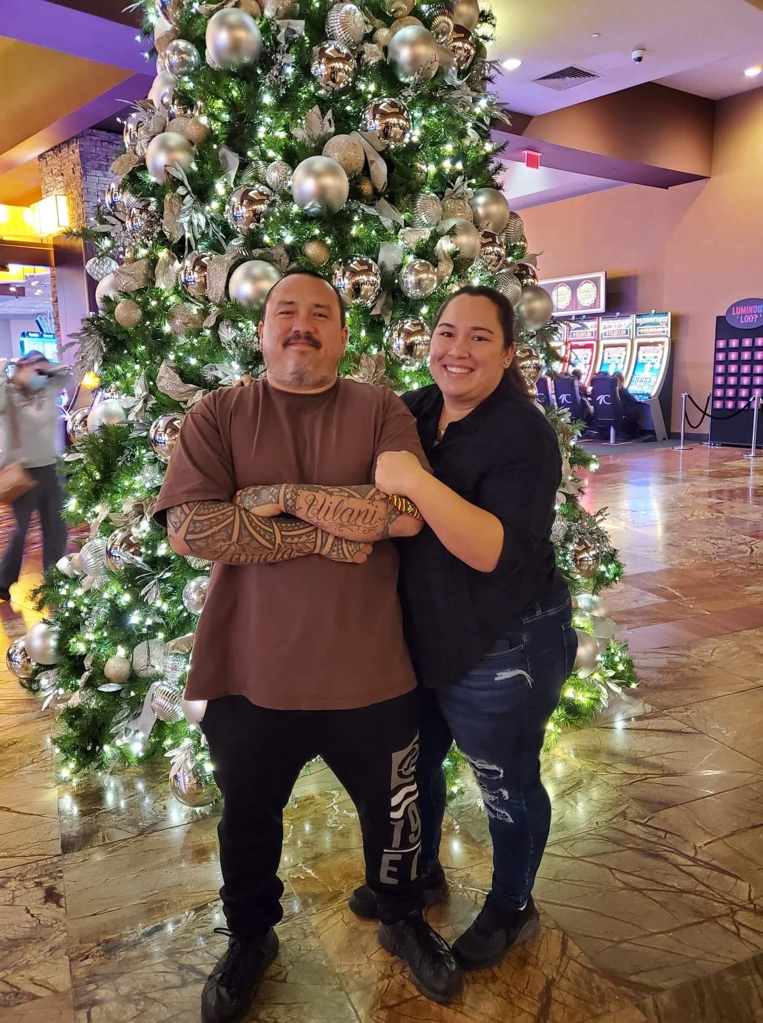 Two people stand in front of a large Christmas tree in a busy area filled with light.