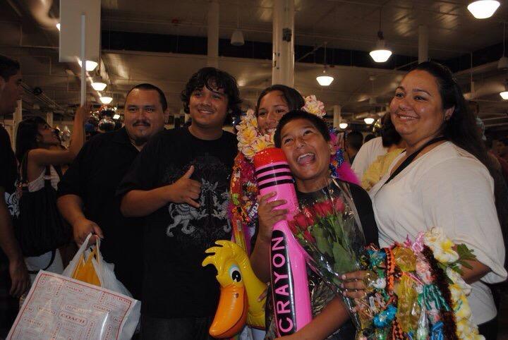 Five people smile at a busy market, holding colorful items together.