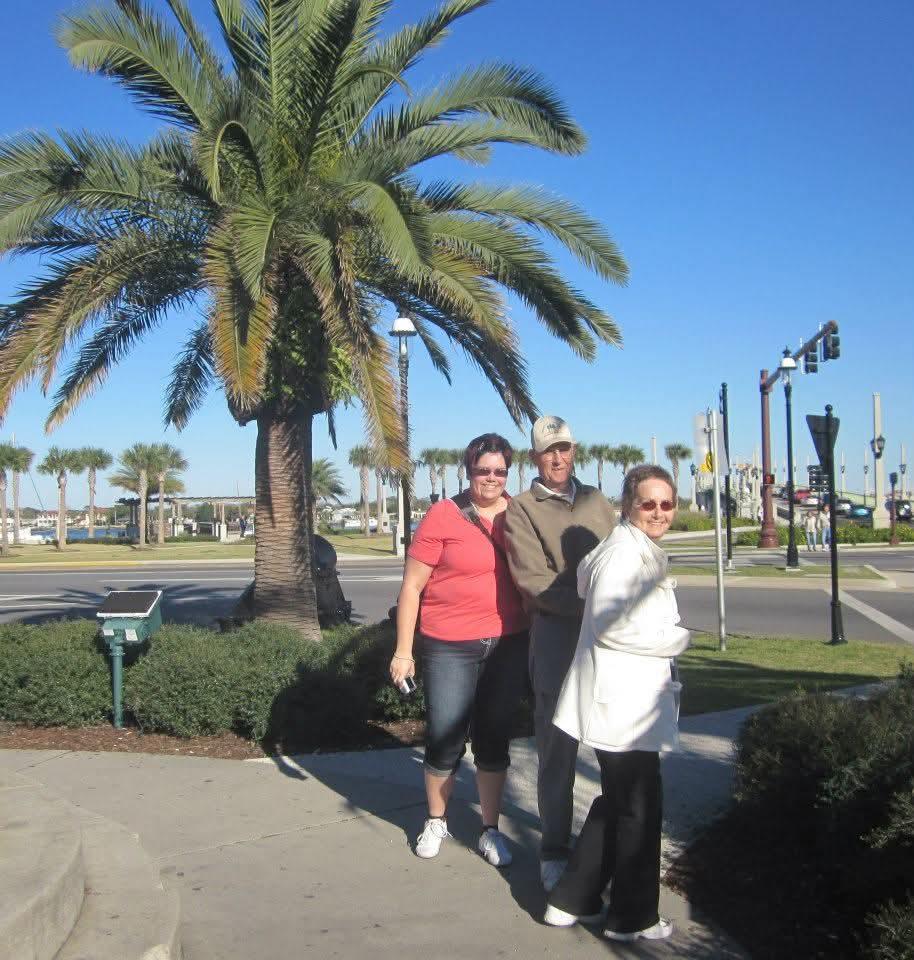 Three friends stand together smiling, outside near a palm tree on a sunny day by the water.