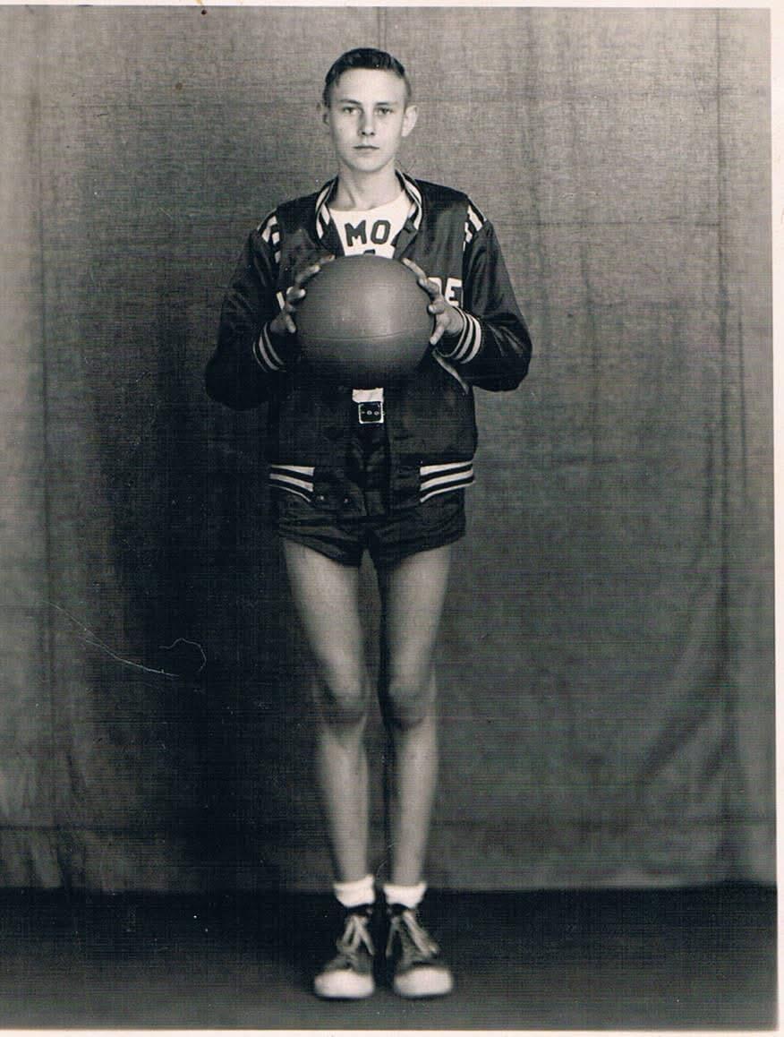 A young person poses with a basketball in a studio. They wear a sports jacket and shorts.