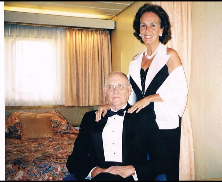 A man sits in a chair while a woman stands behind him in a hotel room. They wear formal clothes.