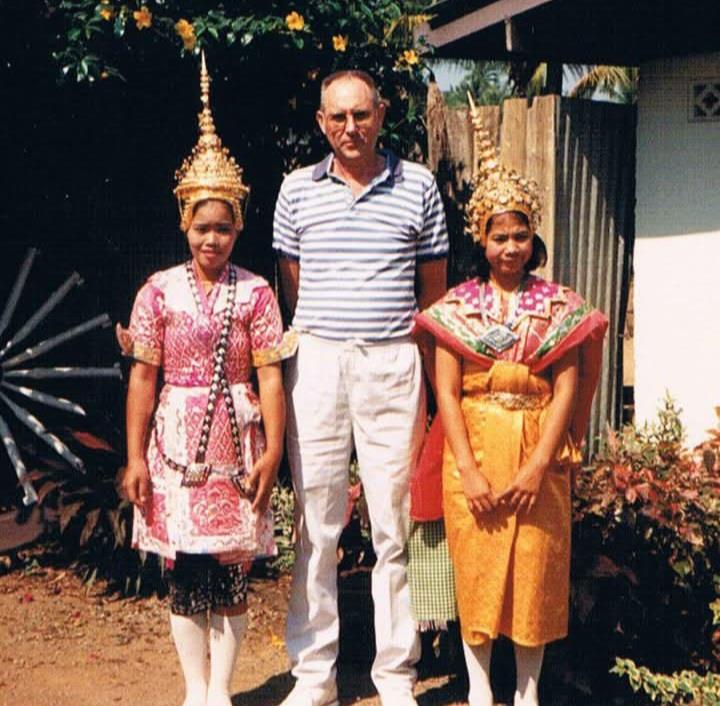 Two women in bright traditional outfits stand with a casually dressed man during the day.