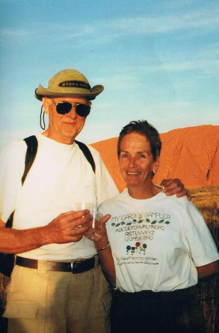 A man and a woman smile as they stand together, holding drinks in the desert near Uluru at sunset.
