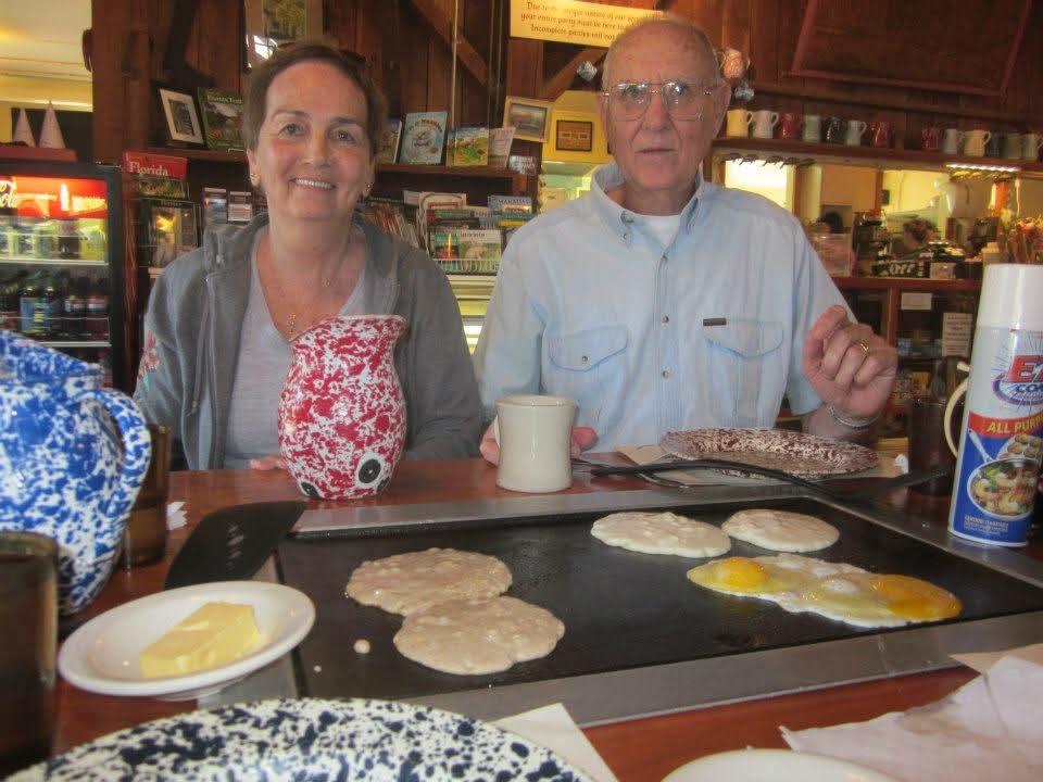 Two people sit at a table with pancakes cooking on a hot grill and drinks in front of them.