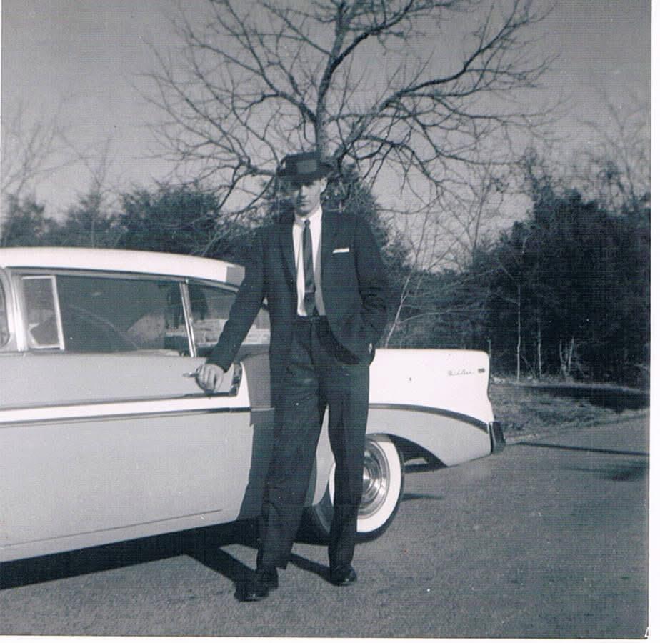 A man in a suit stands beside a vintage car on a dirt road surrounded by trees.
