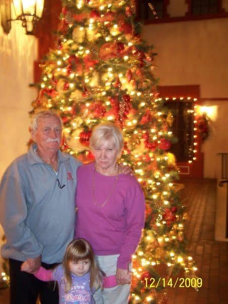 A couple poses with a young girl in front of a large Christmas tree filled with lights.