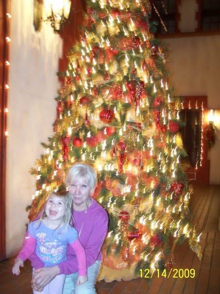 A woman and a girl smile together near a brightly lit Christmas tree during the holidays.