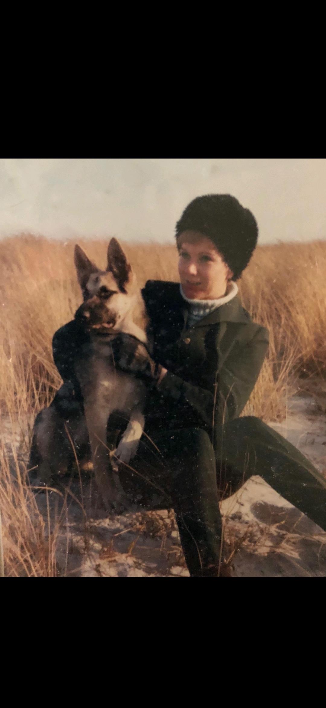 A woman holds a dog while sitting outside in a grassy field under the sun.