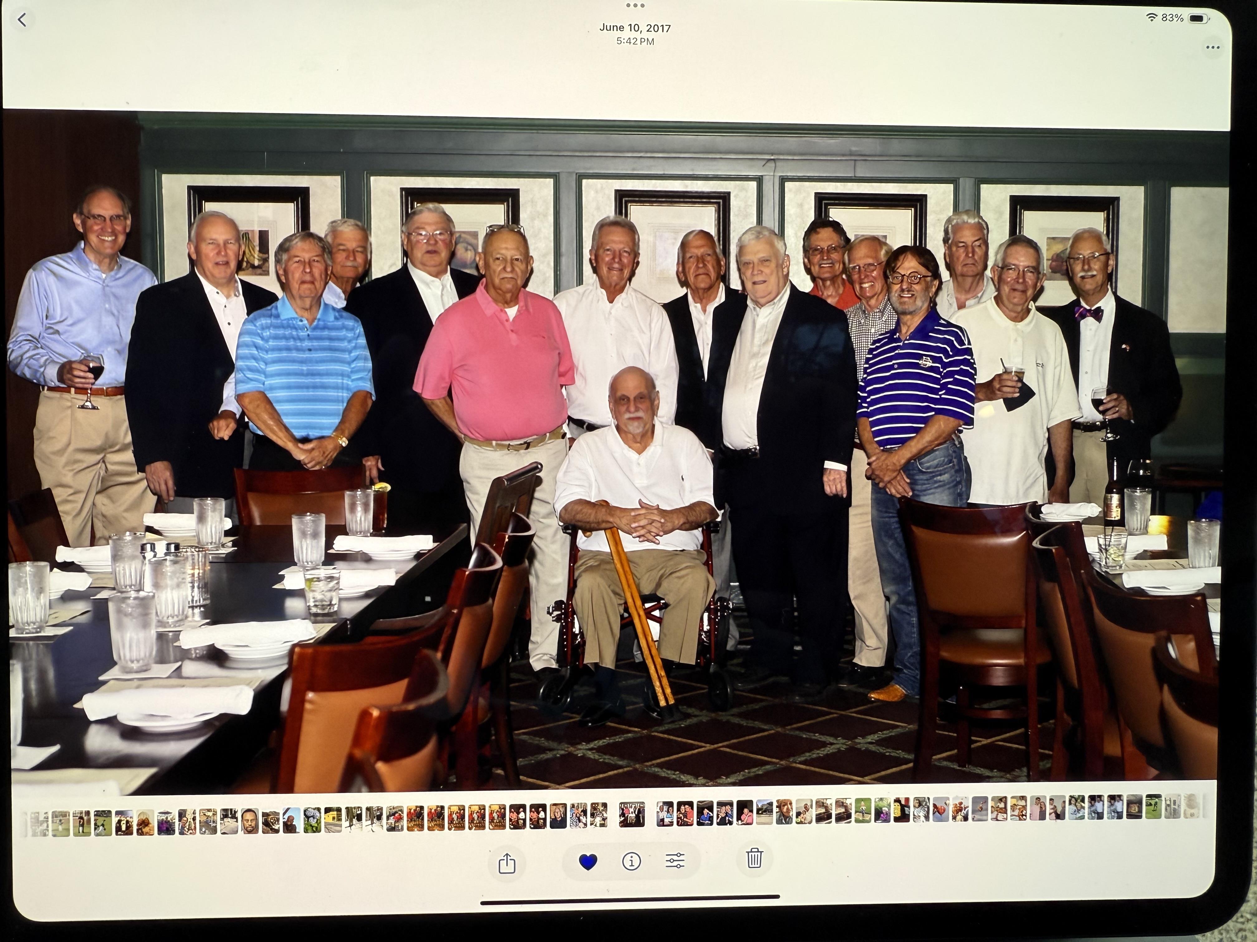 Men pose together at a long table during a meal, all wearing different colors and smiling.