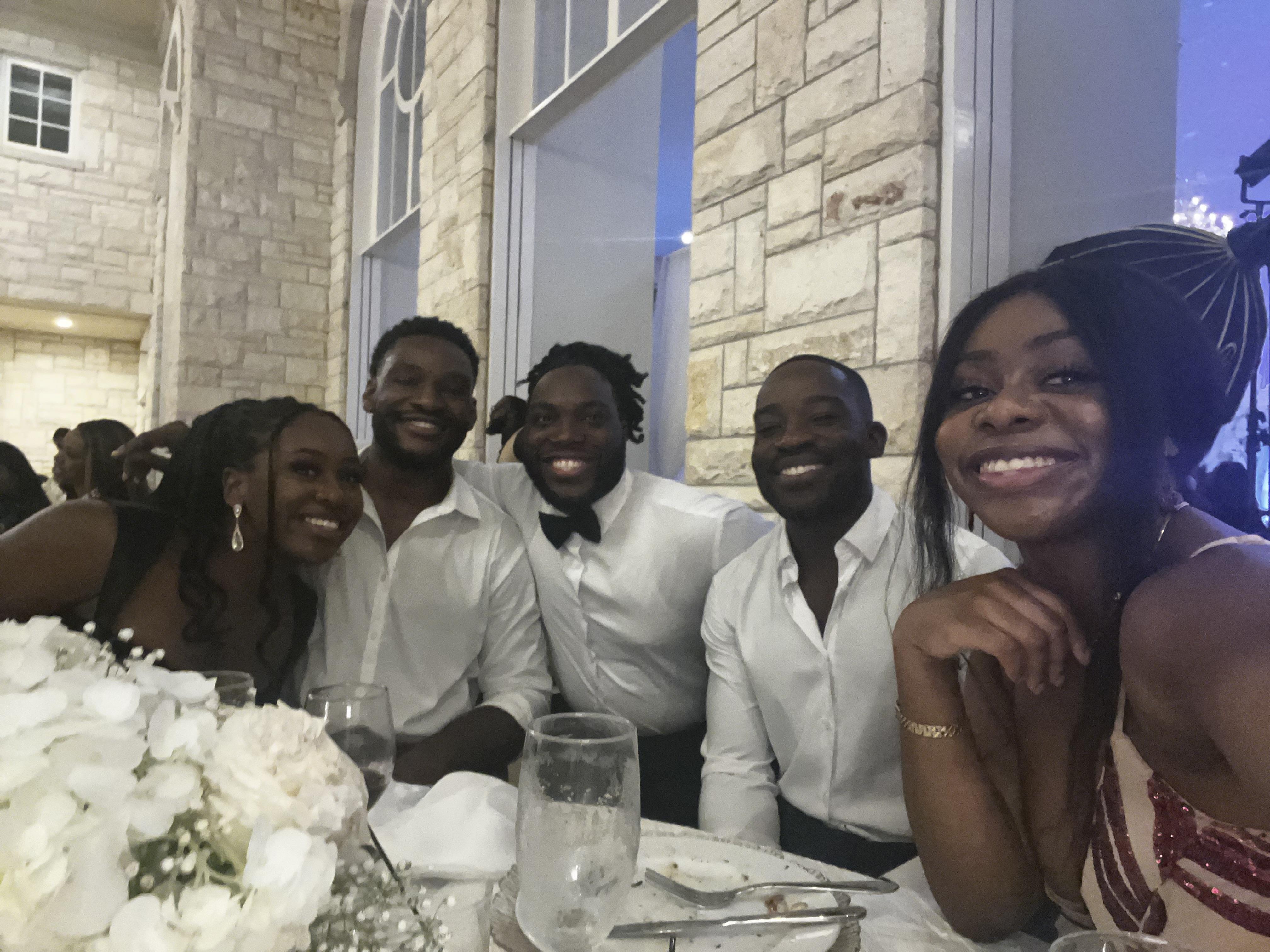 Five friends share laughter and smiles while sitting at a dinner table with drinks and flowers.