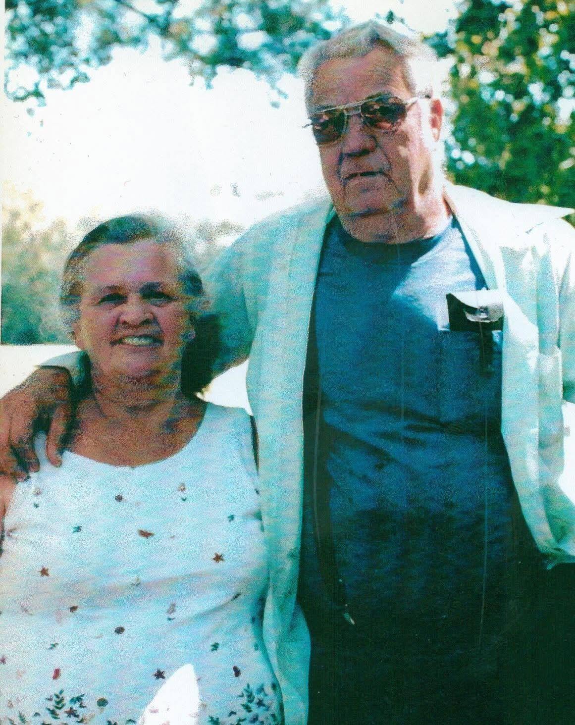 An elderly couple is standing side by side outdoors with sunlight shining through trees.