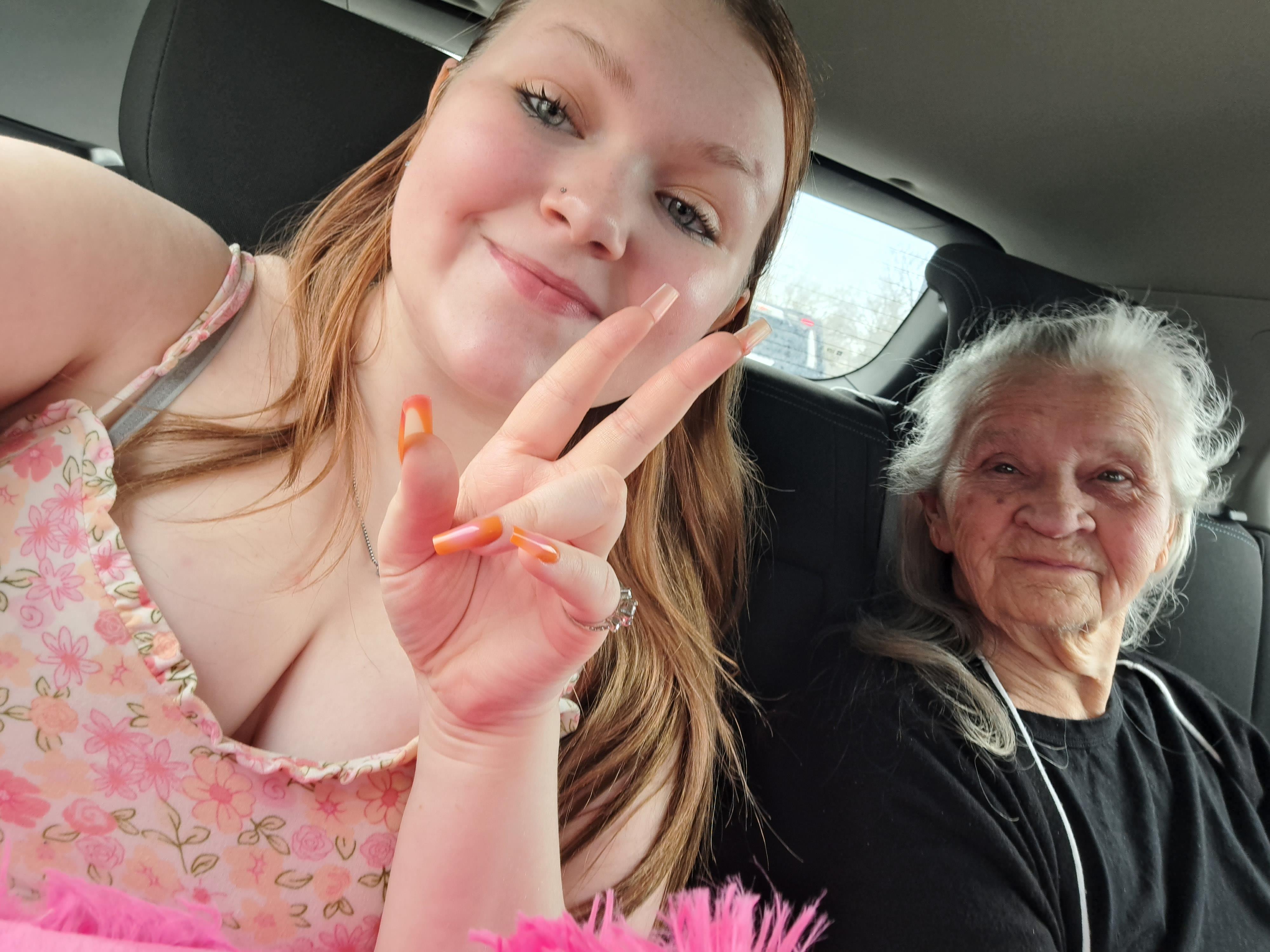 A girl shows a peace sign while sitting next to an older woman in a car. Both seem happy.