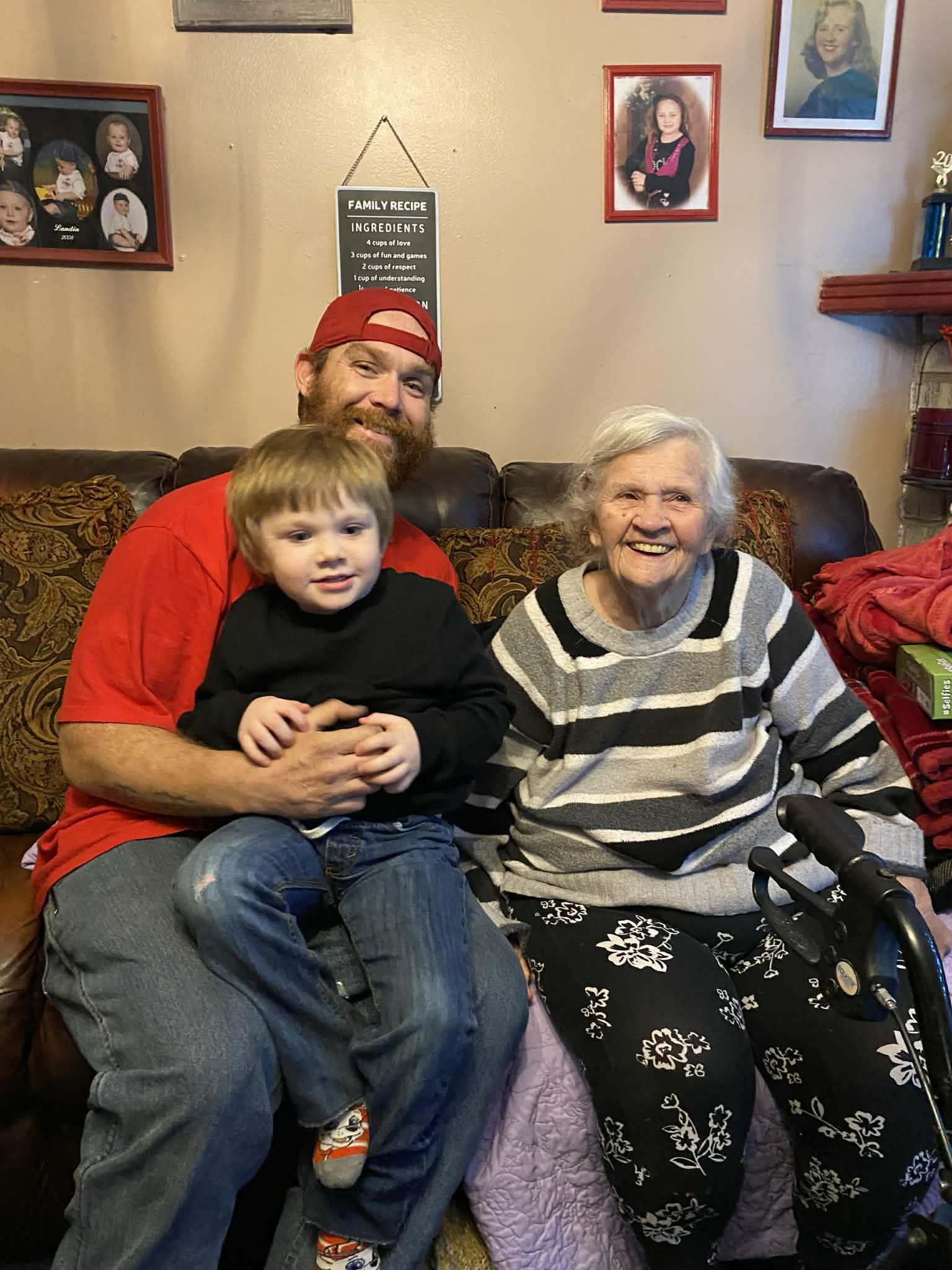 A man holds a young boy while an elderly woman sits nearby, smiling, in a home setting.