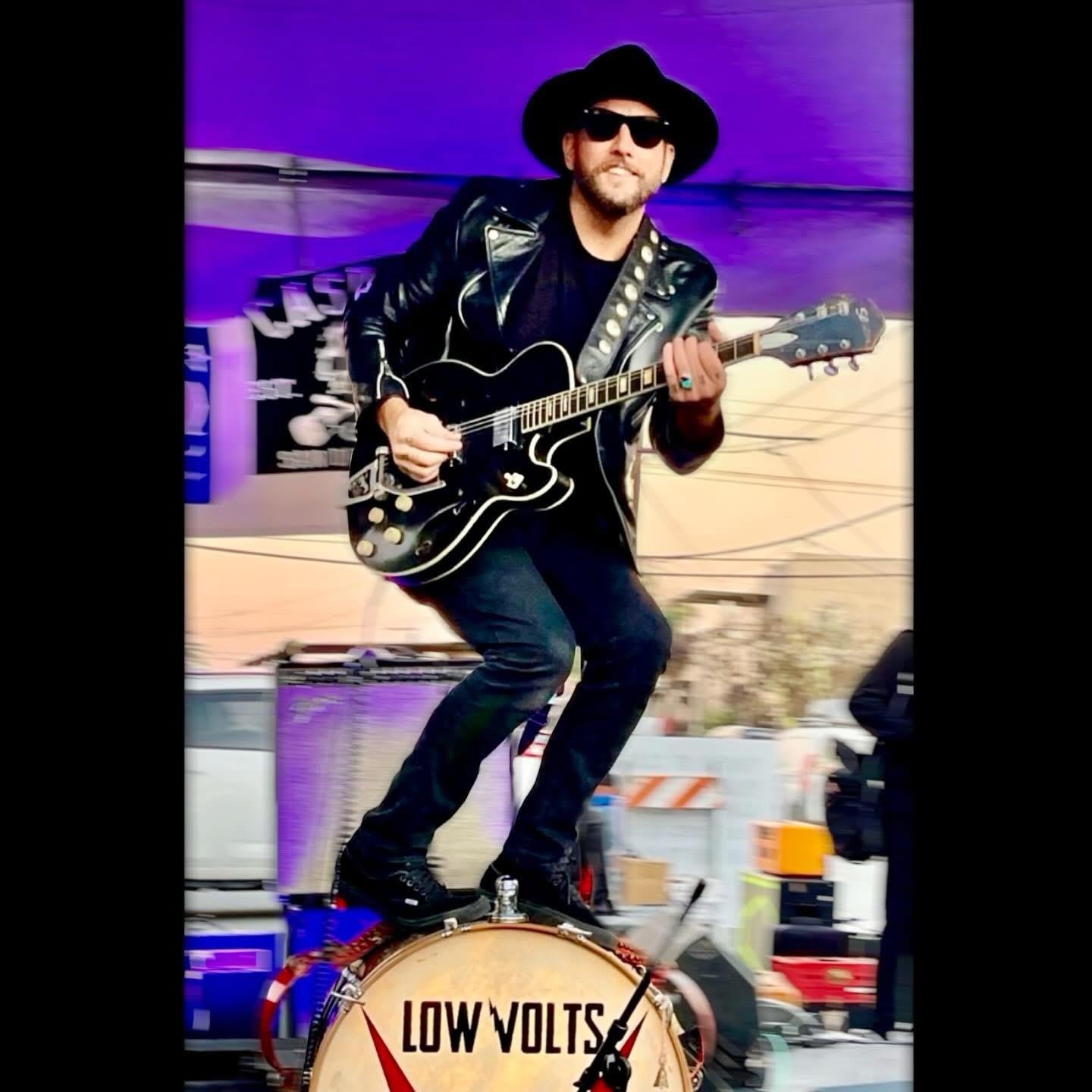 Musician stands on a drum while playing guitar at a concert in an outdoor venue during summer.