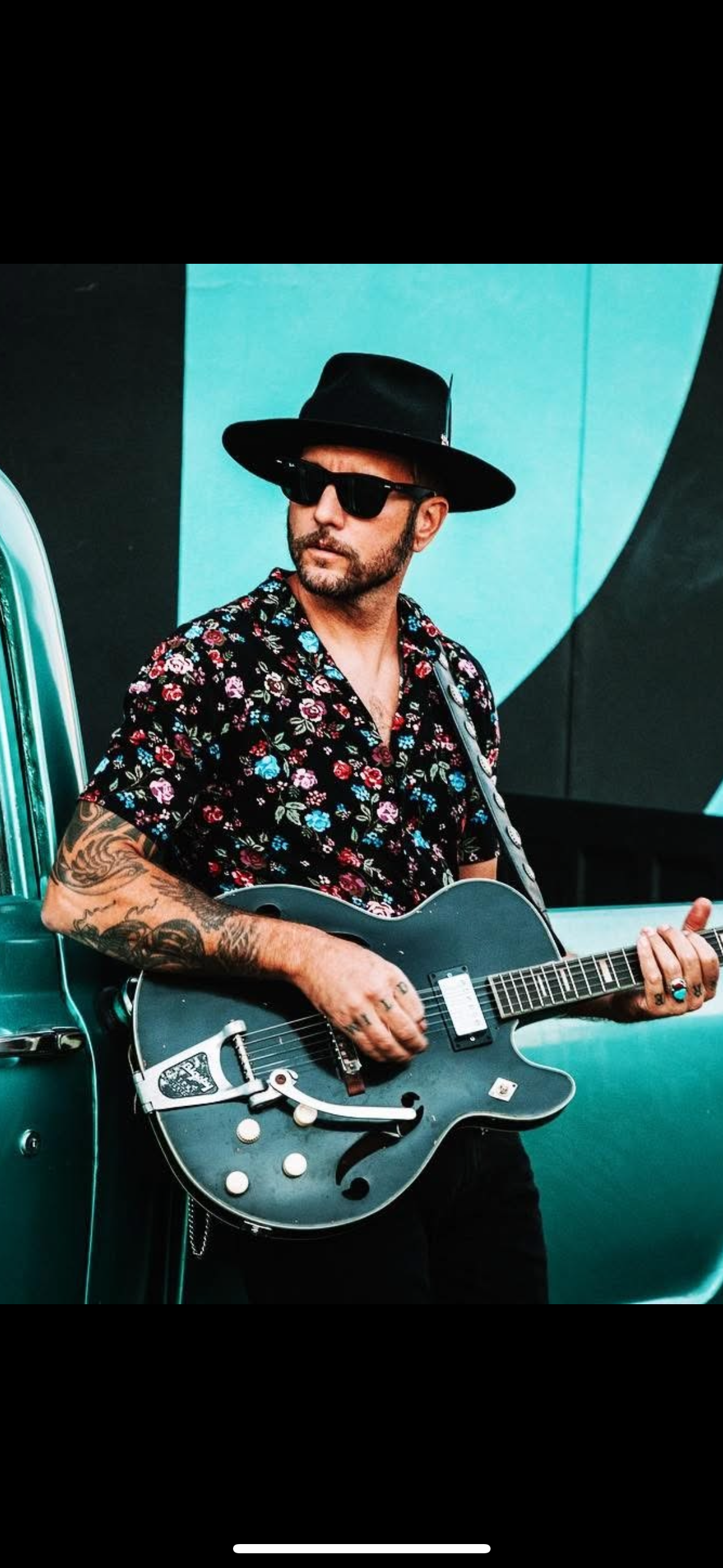A man stands with a guitar in front of a colorful wall during the day.