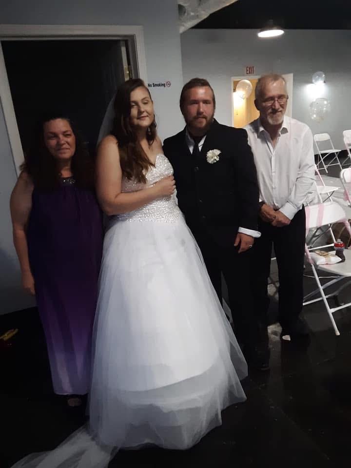 Couple poses with two adults during wedding reception in a lit room with white chairs.