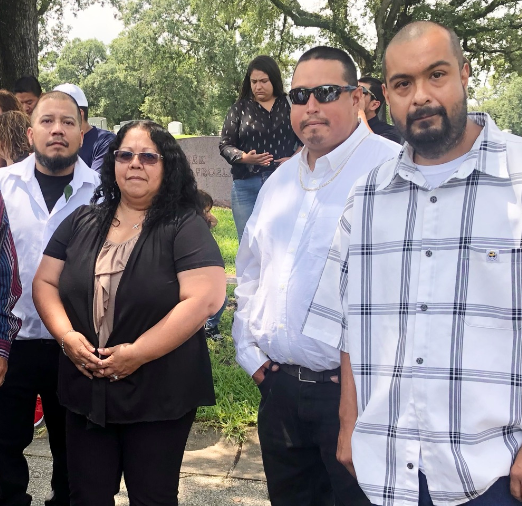 A group of five people stands together in a park during a family gathering near trees.