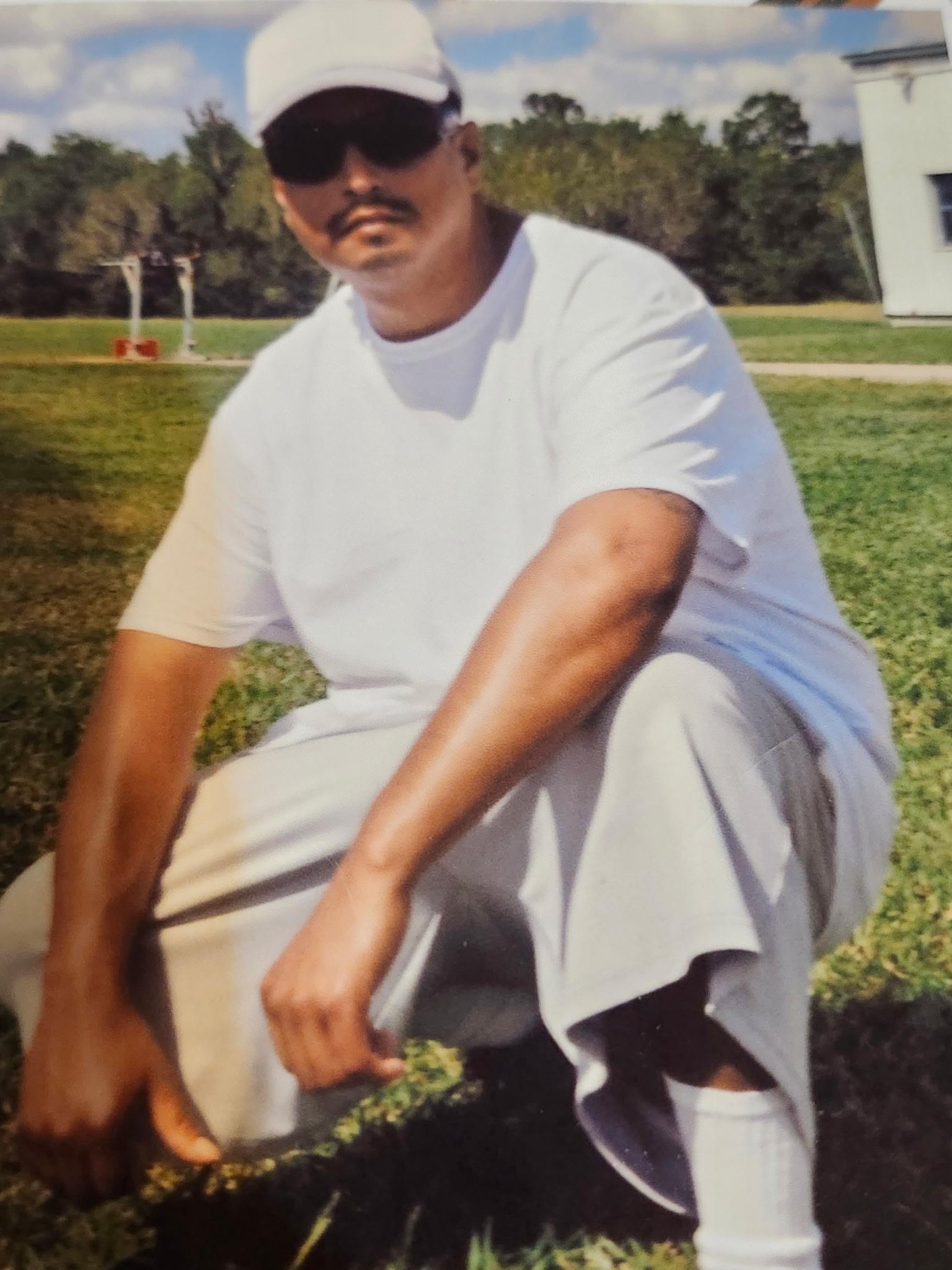 A man in a white shirt squats on green grass in a daytime park, wearing sunglasses.