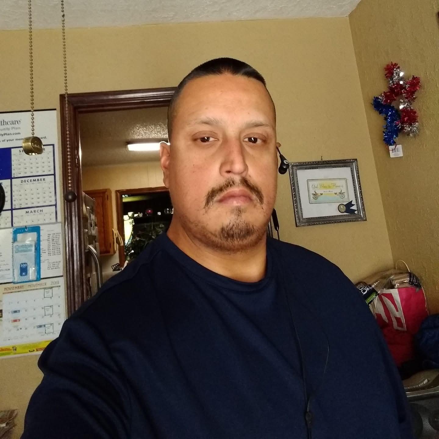 Man stands in a room with basic furniture and wall decor while looking directly at the camera.